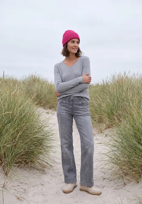 Frau mit pinker Mütze, grauem Pullover und Hose steht auf einem Sandpfad umgeben von Dünengras.