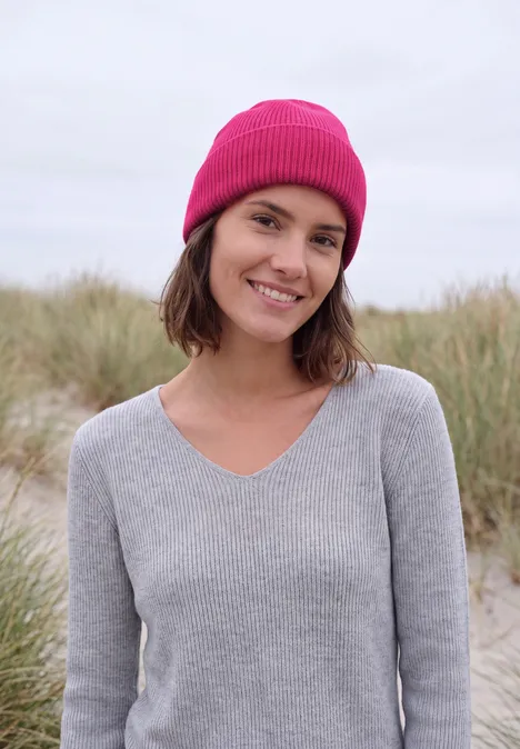 Eine lächelnde Frau mit pinker Mütze und grauem Pullover vor grasbewachsenen Dünen.