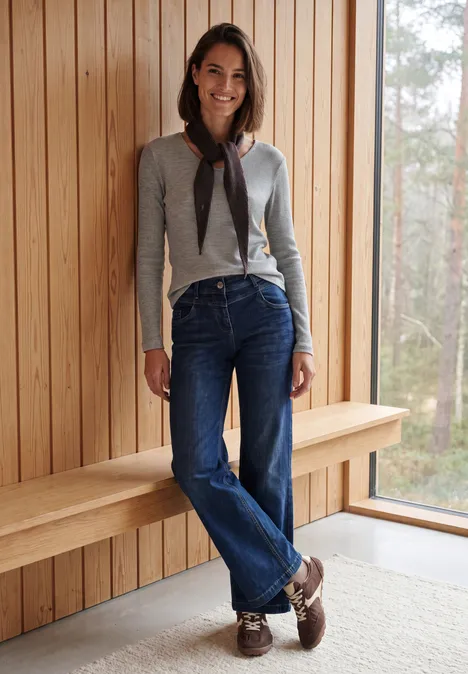 Eine lächelnde Frau lehnt an Holzwand, trägt grauen Cardigan, Jeans und braune Loafers.