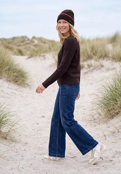 Frau in schwarzem Pullover, Jeans, Beanie und weißen Sneakers spaziert am Dünenstrand.