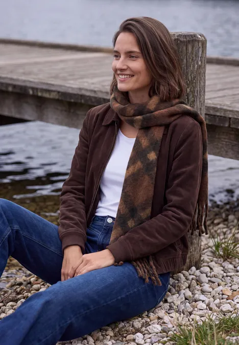 Femme en jean, veste marron et écharpe, assise souriante sur des galets au bord de l'eau.