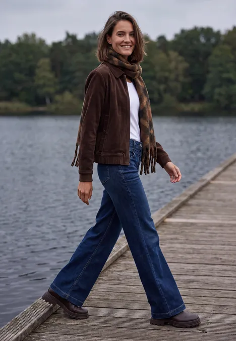 Une femme en veste brune et jean marche en souriant sur une jetée en bois au bord du lac.