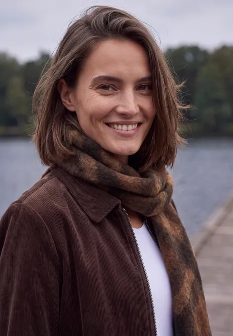 Une femme souriante en manteau marron et écharpe près d'un lac.