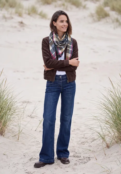 Eine Frau steht in Jeans, Jacke und Schal mit verschränkten Armen lächelnd auf sandigen Dünen.