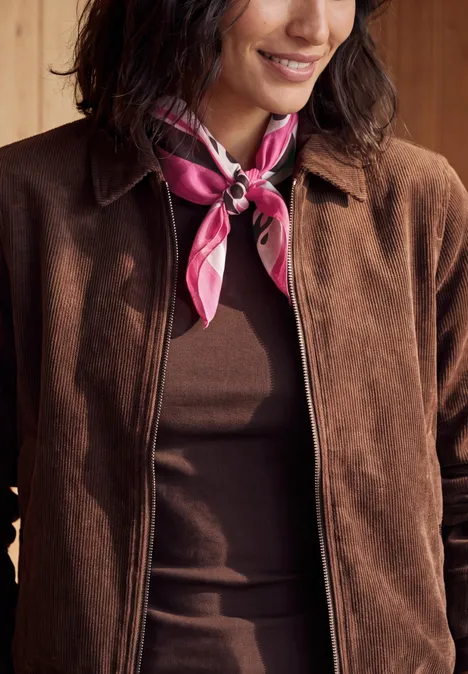 Personne en veste marron et top assorti, portant un foulard rose noué autour du cou.