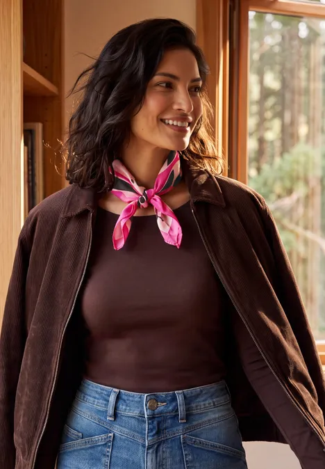 Femme appuyée sur une étagère, pull marron, jean, foulard rose, souriante.