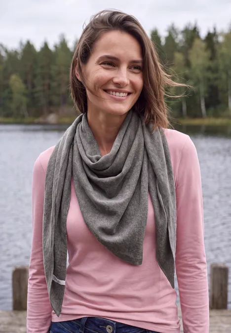 Lachende vrouw met roze trui en grijze driehoekssjaal bij een bosachtig meer.
