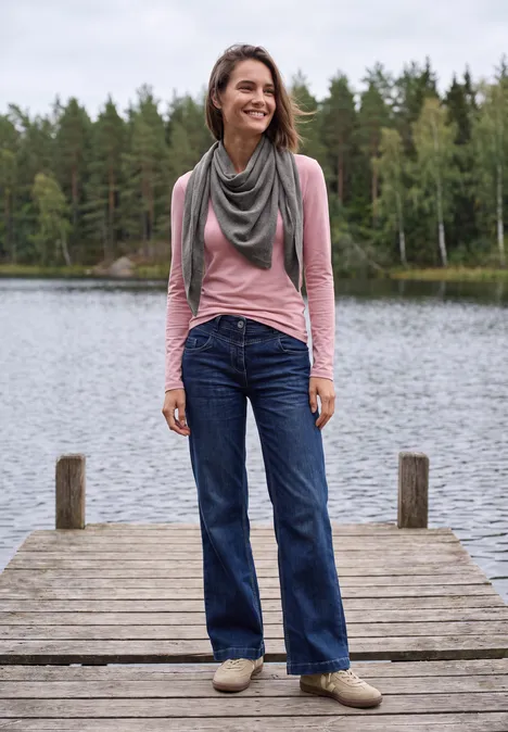 Vrouw lacht op een steiger bij een meer, draagt spijkerbroek, roze shirt en sjaal, bos op de achtergrond.