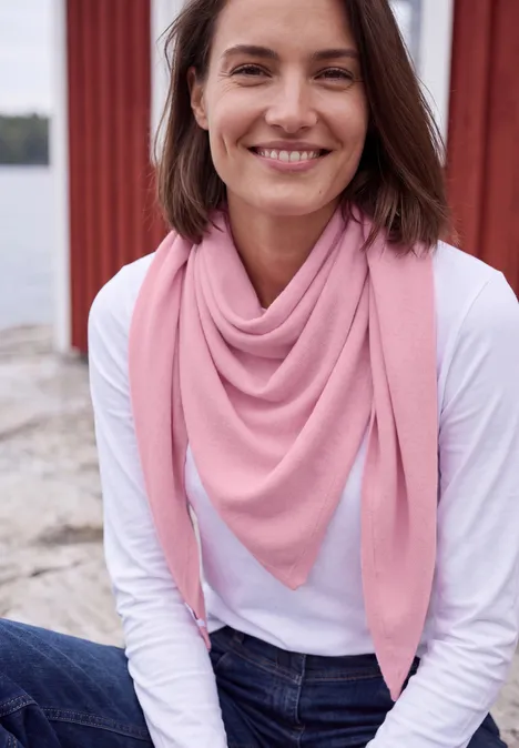 Een glimlachende vrouw in een wit shirt en een roze driehoekige sjaal zit aan het water.