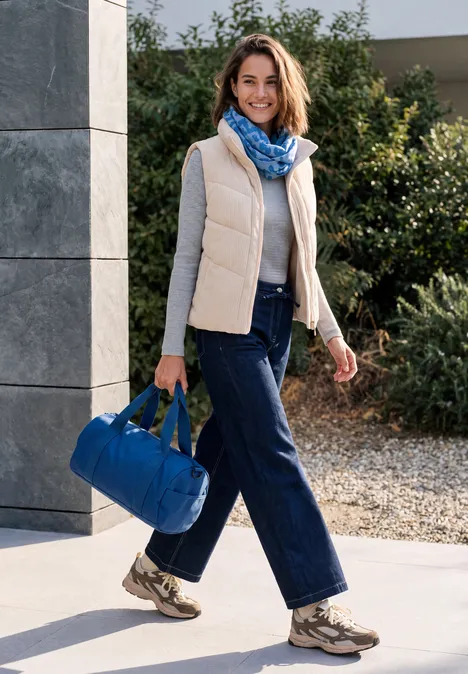 Frau in Freizeitkleidung mit einer blauen Reisetasche, lächelnd vor einem modernen Haus.