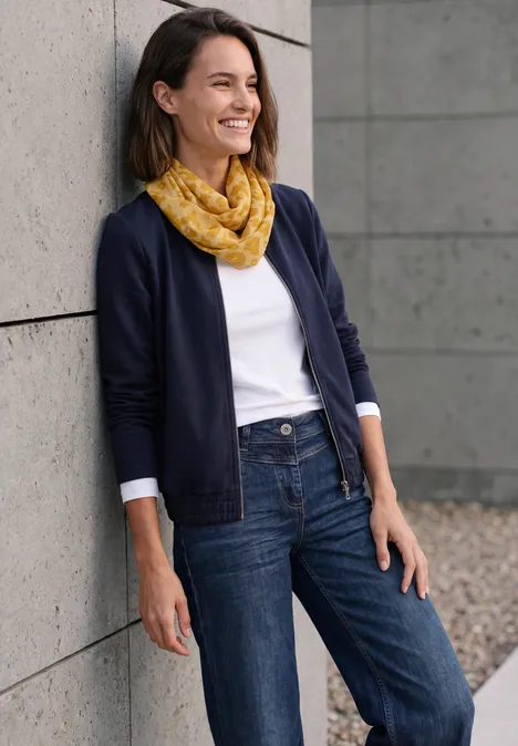 Eine lächelnde Frau lehnt im Freien an einer Wand, trägt Jeans, weißes Shirt, dunkelblauen Cardigan und gelben Schal.