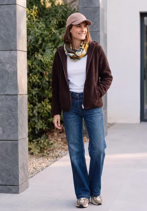 Eine lächelnde Frau in Jeans, weißem T-Shirt, brauner Jacke und Schal steht vor einem Haus und schaut zur Seite.