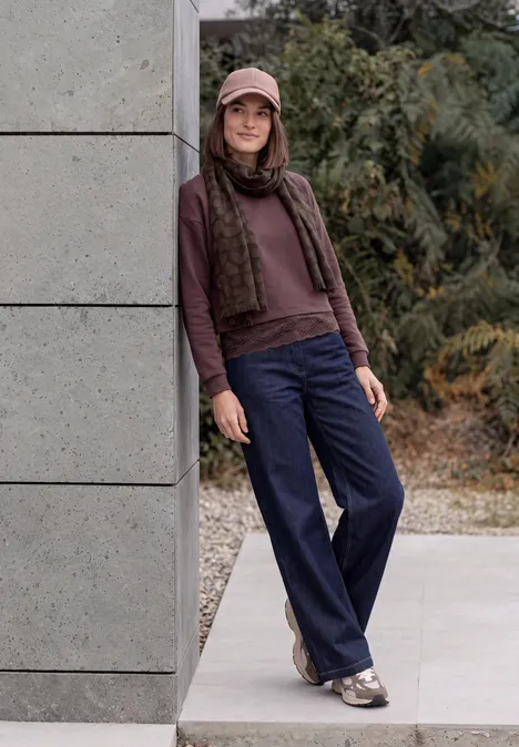 Eine junge Frau lehnt draußen an einer Wand, trägt Mütze, Schal, Pullover, blaue Hosen und Sneaker.