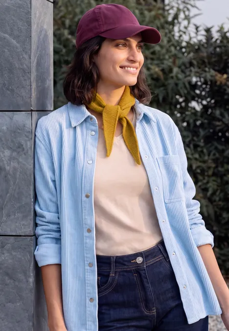 Schmaler Schal in Unifarbe golden honey melange Frau mit violettem Cap, gelbem Halstuch, beigem Pullover und blauem Hemd lächelt draußen neben einer Mauer.