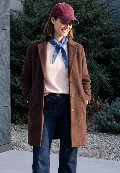 Vrouw met rode pet, bruine jas, spijkerbroek en blauwe sjaal, buiten glimlachend.