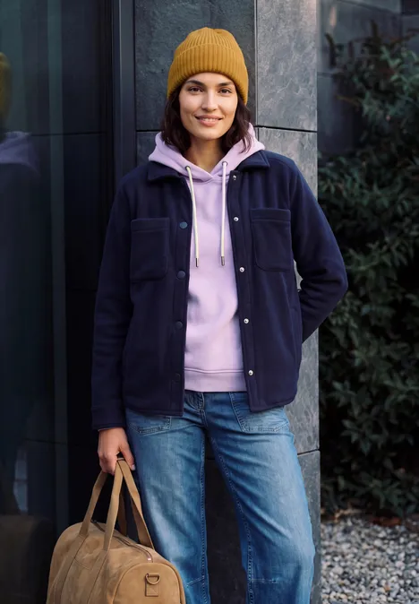 Eine junge Frau steht draußen vor einem Gebäude, trägt eine gelbe Mütze, rosa Hoodie, Jeans und hält eine Tasche.