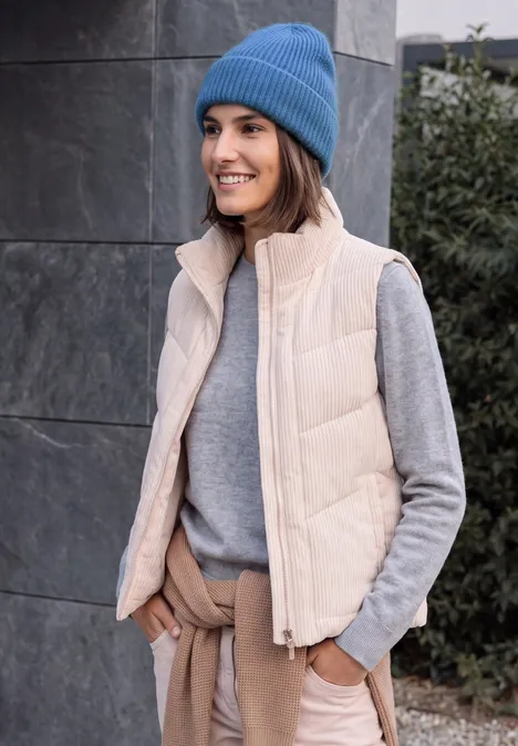 Vrouw met blauwe muts, grijze trui en beige bodywarmer lacht met handen in de zakken.