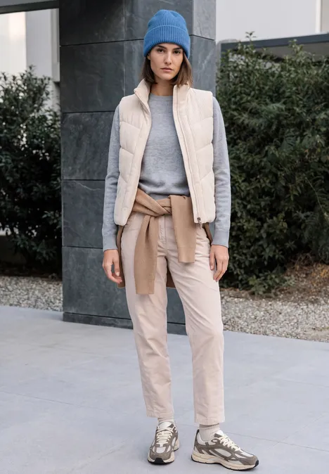 Vrouw in comfortabele buitenkleding: blauwe muts, grijze trui, beige bodywarmer en broek, sneakers.