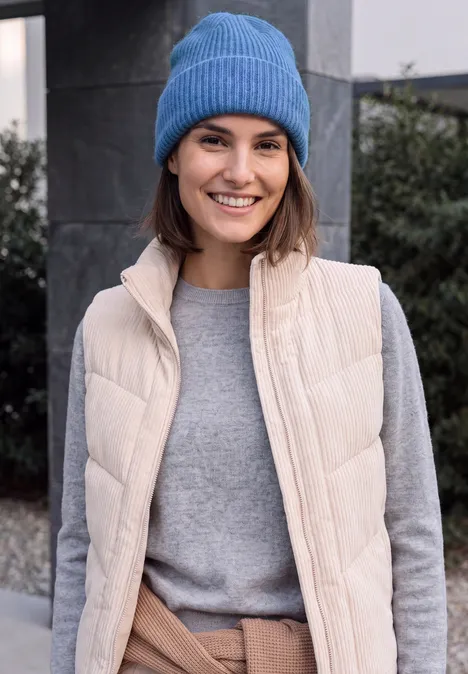 Een glimlachende vrouw met een blauwe muts, beige bodywarmer en grijze trui buiten.
