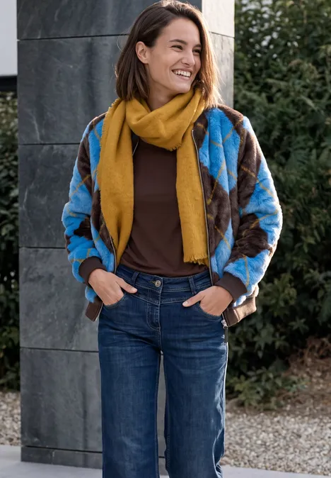 Eine lächelnde Frau in Jeans, blau-brauner Jacke und gelbem Schal steht entspannt an einer Säule im Freien.