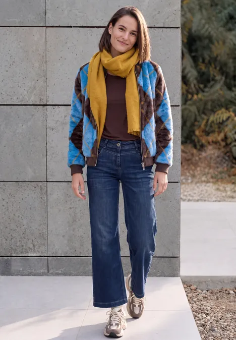 Eine Frau steht vor einer Betonwand, trägt bunte Patchworkjacke, gelben Schal, Jeans und Halbschuhe.