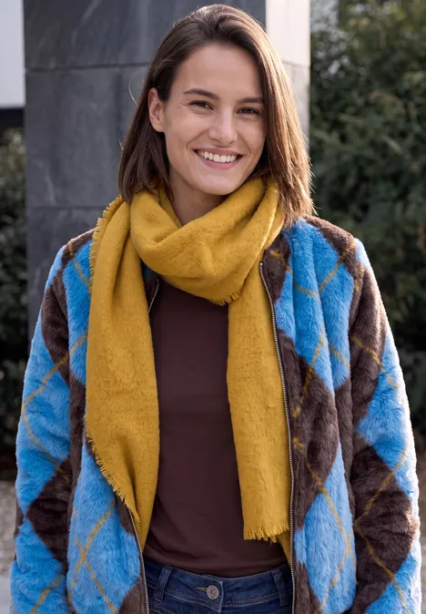 Eine lächelnde Frau mit blauer, brauner Rautenstrickjacke und gelbem Schal im Freien.