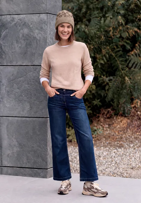 Junge Frau in Beanie, hellem Pullover, weiten Jeans und Sneakers steht vor einer Steinwand und grüner Kulisse.