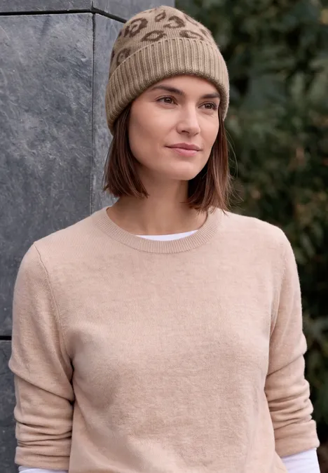 Junge Frau im beigen Pullover und gestrickter Mütze steht vor einer Steinwand, dahinter grüne Bäume.