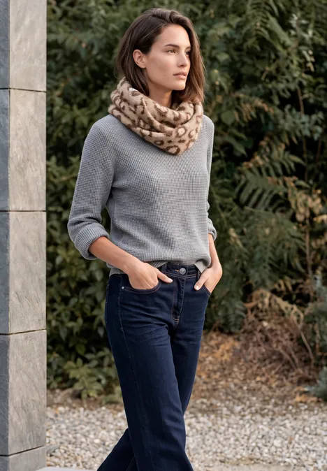 Frau in grauem Pullover und Schal, dunkle Jeans, Hände in Taschen, steht draußen an Säule.