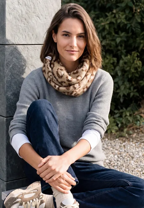 Eine Frau sitzt im Freien auf Kies, trägt grauen Pullover, Schal mit Leopardenmuster und Jeans.