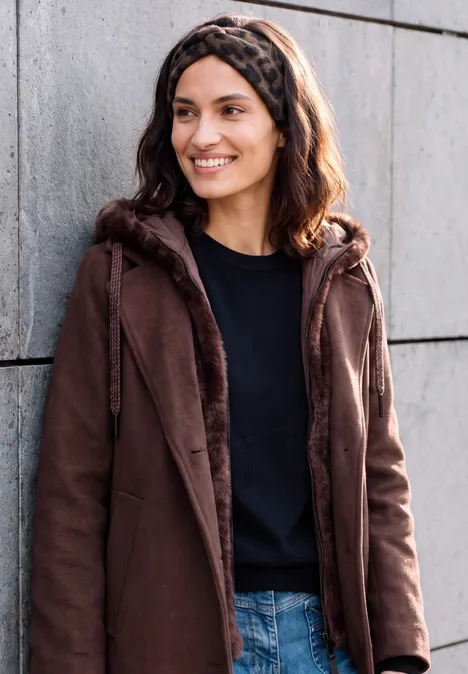 Een glimlachende vrouw in een bruine jas, zwart shirt en jeans leunt tegen een grijze muur.