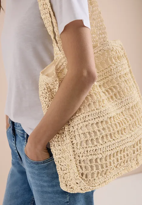 Frau in weißem T-Shirt und Jeans trägt an Arm eine gehäkelte, beige Tasche.