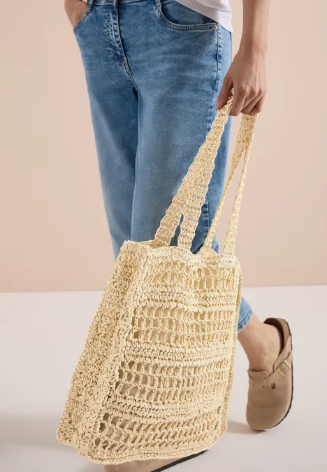 Eine gehäkelte Einkaufstasche aus Naturgarn hängt neben Jeans und beigen Hausschuhen.