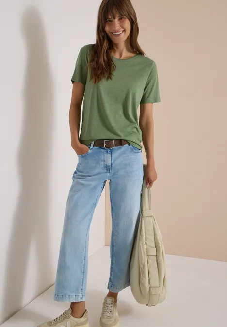 Basic riem met vierkante gesp Cacao Frau lehnt an Wand, trägt grünes T-Shirt, weite blaue Jeans mit Gürtel und hält lässige Tasche.