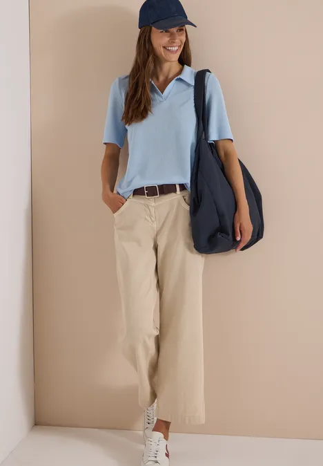 Basic riem met vierkante gesp Raisin Frau trägt hellblaues Poloshirt, beige Stoffhose, weiße Sneaker, blaue Cap und große Umhängetasche.