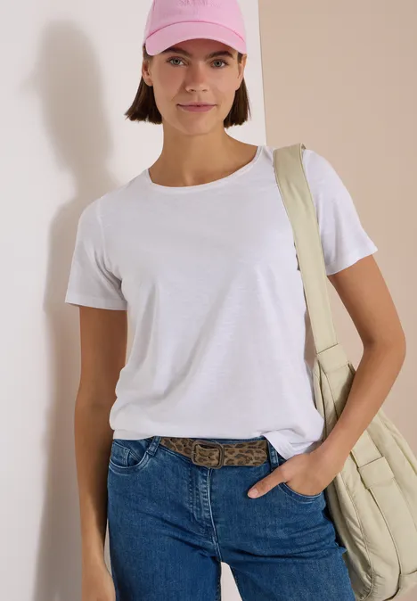 Gürtel mit Leo-Muster Taupe Leo Frau in weißem T-Shirt, Jeans und rosa Cap mit Umhängetasche, stehend vor heller Wand.