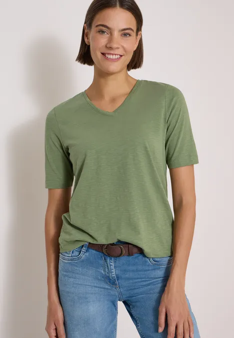 Gürtel mit Streifenmuster Suede Brown Frau mit grünem V-Neck T-Shirt und Jeans lächelt vor hellgrauem Hintergrund.