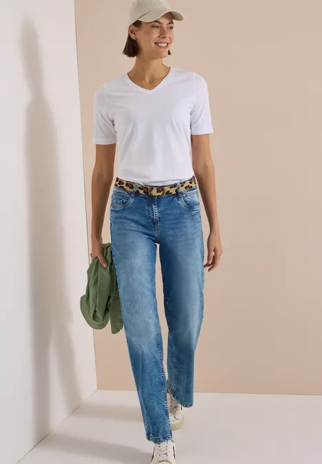Frau in weißem T-Shirt, Jeans und Cap läuft lächelnd, hält grüne Jacke und trägt Leopardenmuster-Gürtel.