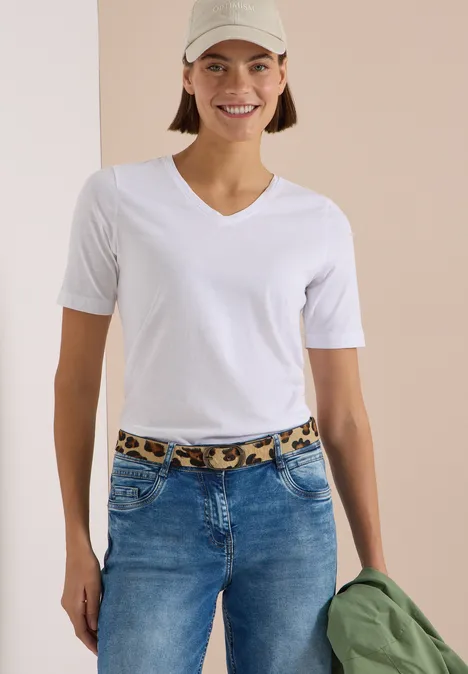 Frau in weißem V-Ausschnitt-T-Shirt und Jeans mit Leoparden-Gürtel, trägt Cap und hält grüne Jacke.