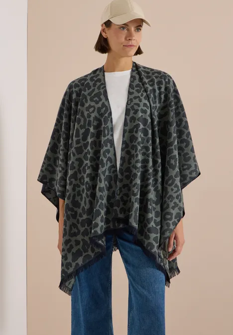 Cape mit Leo-Muster dried olive green Frau mit gemustertem Poncho-Cardigan, weißem T-Shirt, Jeans und Baseballcap vor hellem Hintergrund.