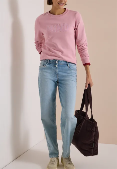 Femme en sweat-shirt rose et jean bleu clair portant un grand sac sombre devant un fond neutre.