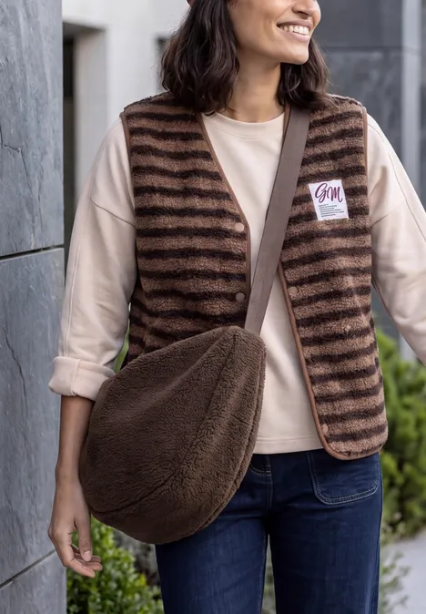 Umhängetasche chocolate sauce brown Frau trägt braun gestreifte Weste, helles Shirt, Jeans und braunen Fell-Umhängebeutel, steht lächelnd draußen.