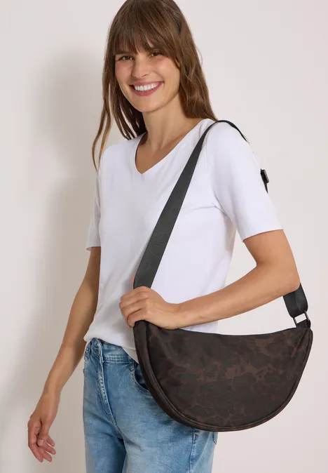 Frau in weißem T-Shirt und Jeans trägt braune Halbmond-Umhängetasche und lächelt.