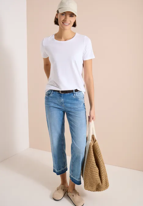 Frau trägt weißes T-Shirt, blaue Jeans, beige Slipper und hält eine große Korbtasche.