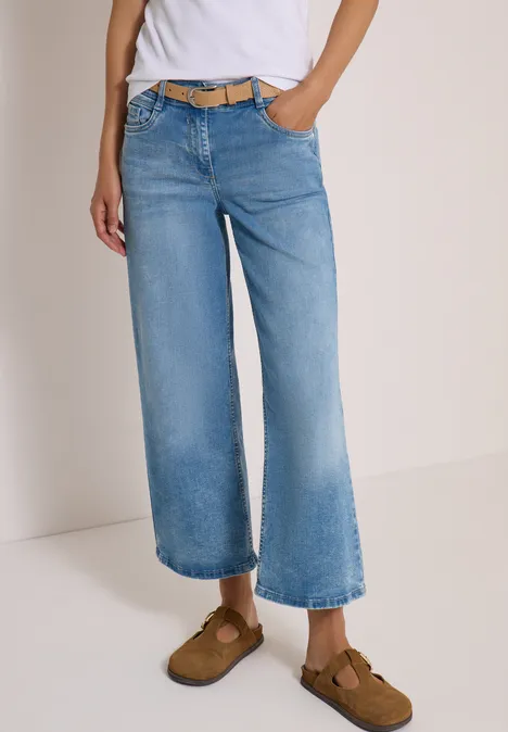 Persoon draagt lichtblauwe, wijdvallende culotte-jeans met riem en bruine loafers, neutrale achtergrond.