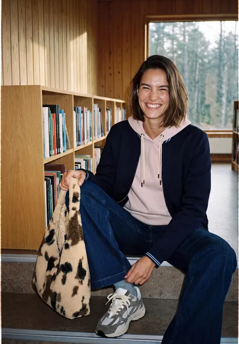 Eine lächelnde Frau sitzt in lockerer Kleidung auf Lesetreppen neben Bücherregalen und hält eine gemusterte Tasche.