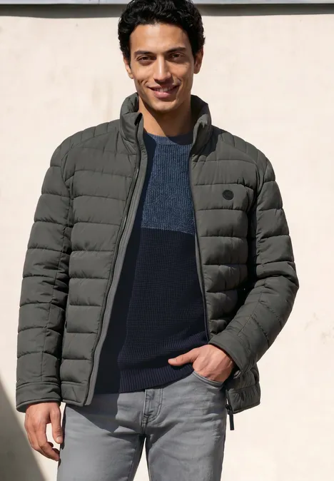 Ein junger Mann steht lässig in einer grauen Steppjacke über dunkelblauem Pullover und Jeans.