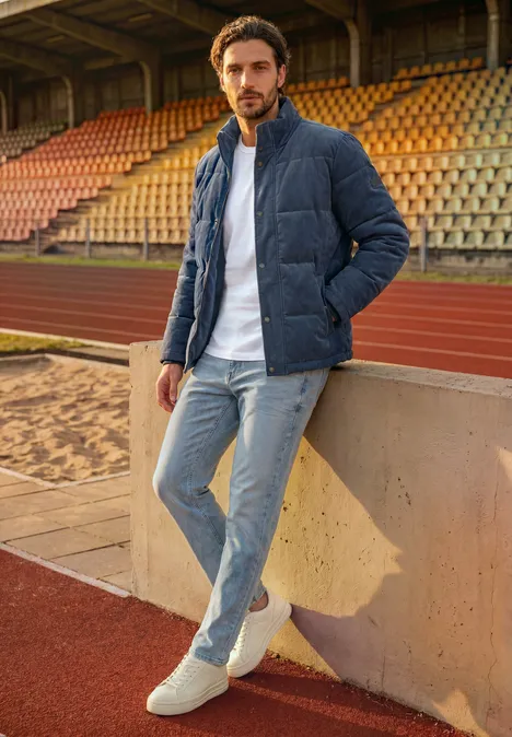 Ein Mann lehnt sich in blauer Steppjacke und Jeans an eine Mauer in einem leeren Stadion mit Laufbahn.