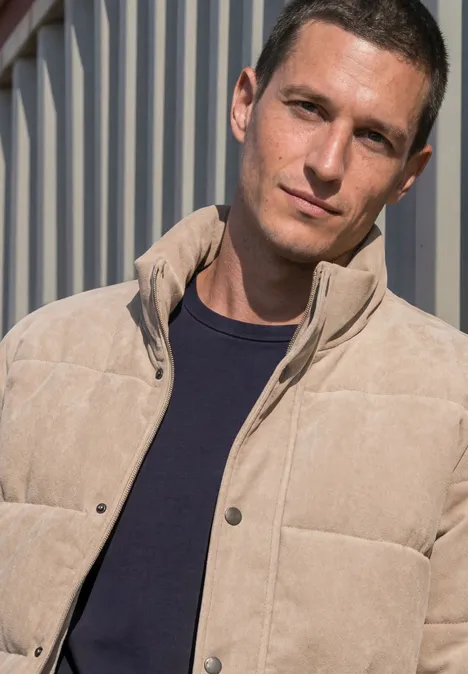 Ein Mann in einer hellen Steppjacke lehnt vor einer Metallwand.