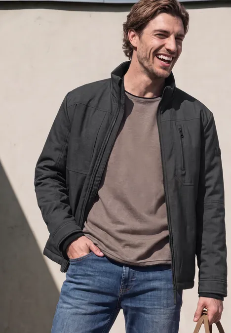 Veste à col montant et fermeture zip ash grey melange Ein lächelnder Mann in schwarzer Jacke, grauem Shirt und blauen Jeans.